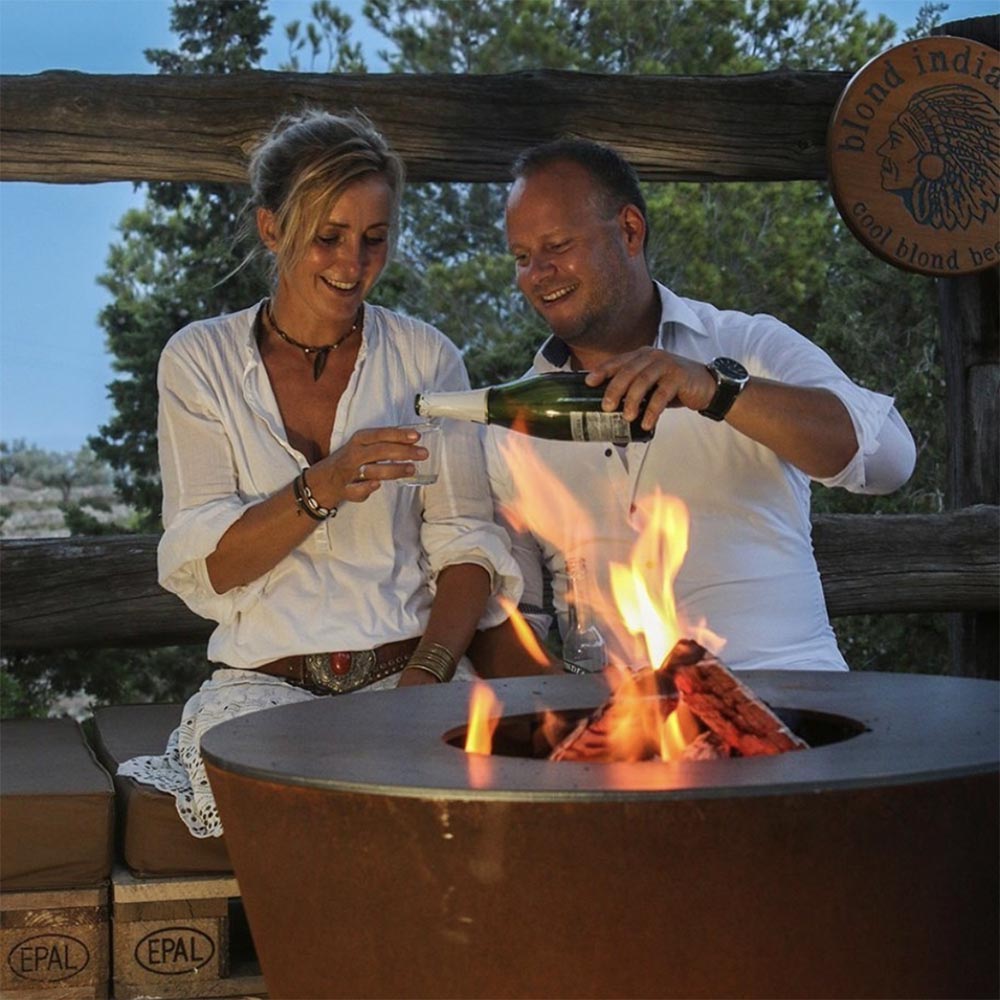 Brown fire pit grill with tall flames, outdoor guests cooking