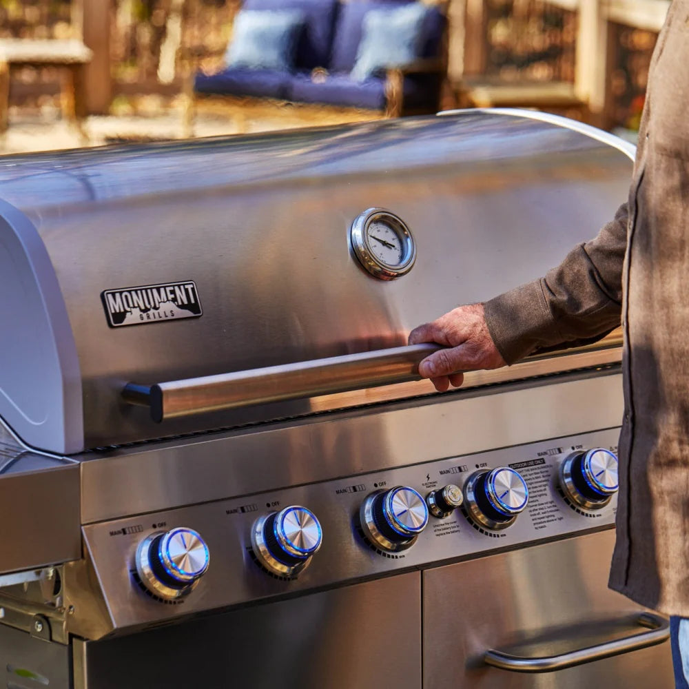 Stainless grill control panel with six blue knobs and 'Monument' lid