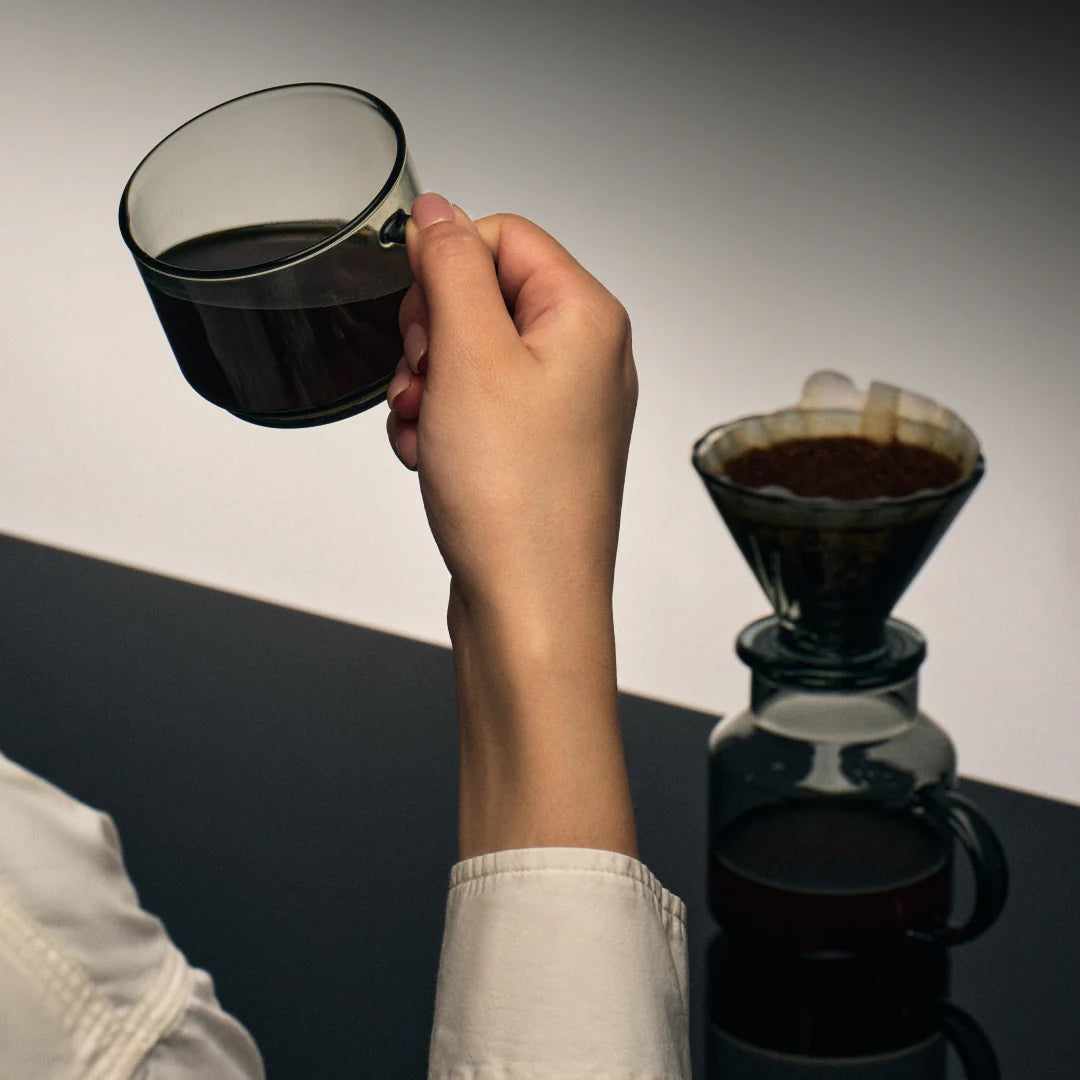 Hand holding a black glass mug next to a pour-over dripper and server
