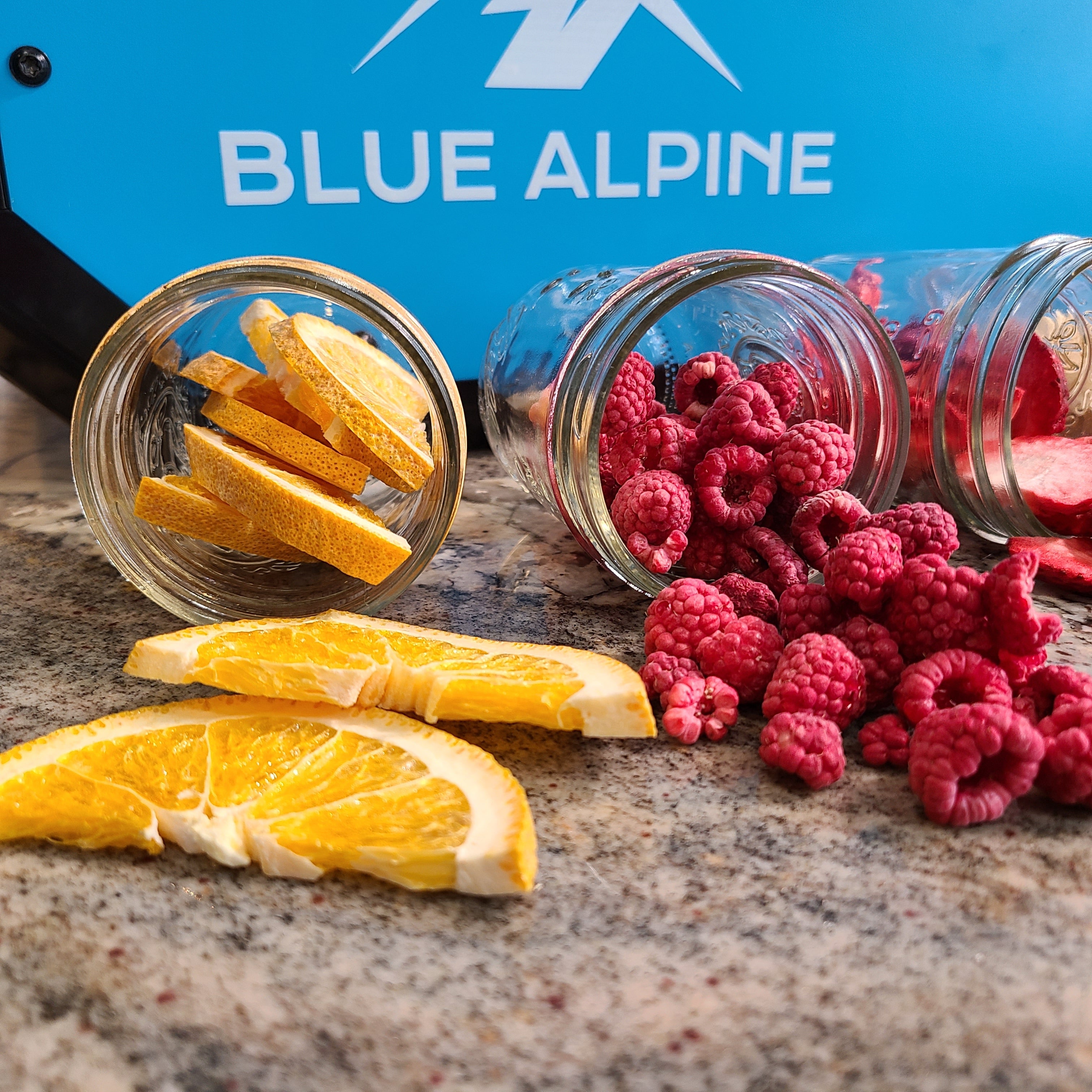 Freeze dryer serving jars with dried orange slices and raspberries, near open unit.