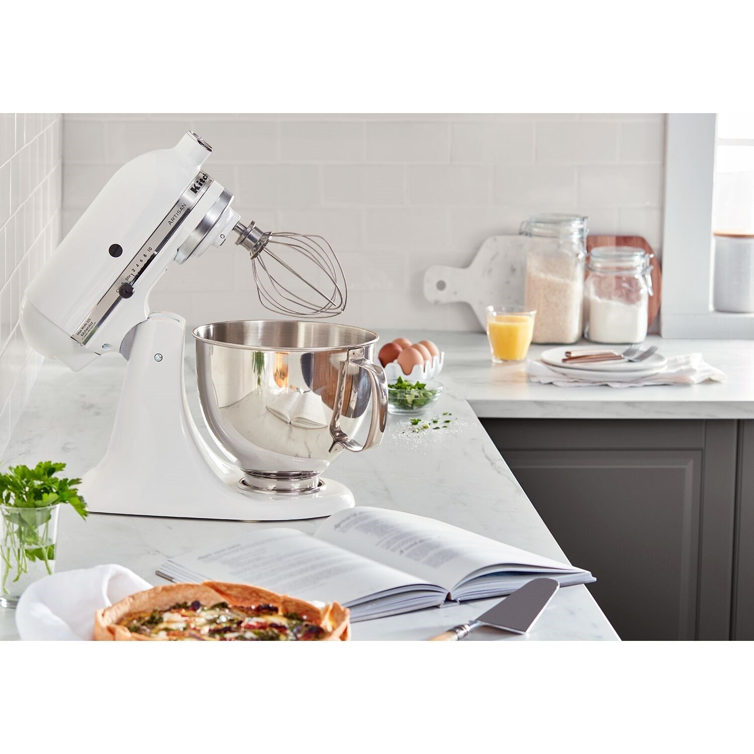 White tilt-head stand mixer with stainless bowl and whisk attachment near ingredients.