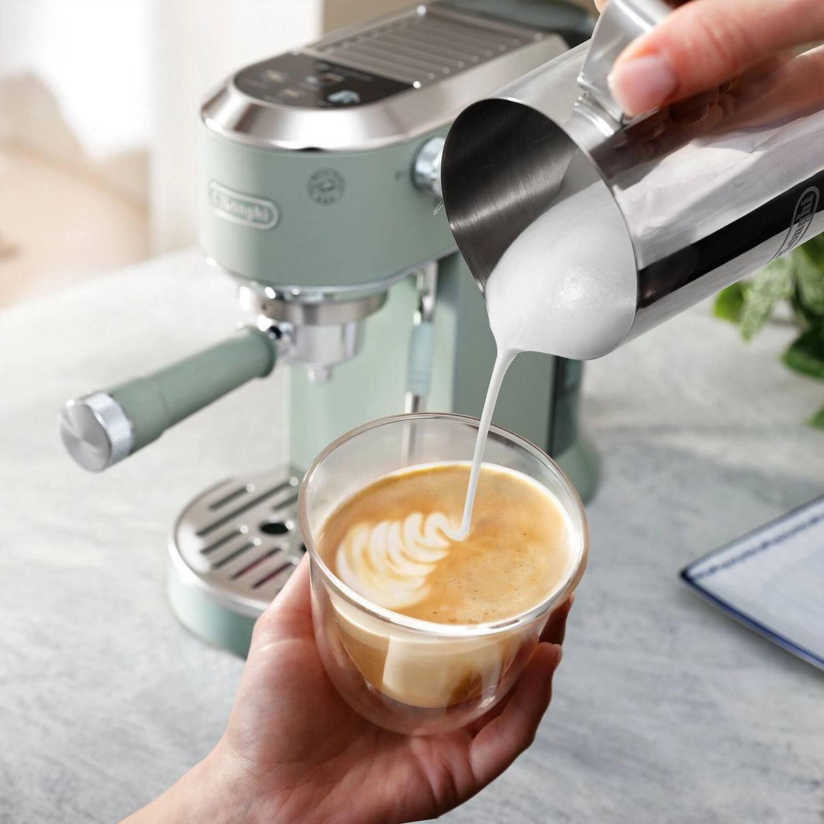Green De'Longhi steam wand pouring into glass cup with creamy espresso foam