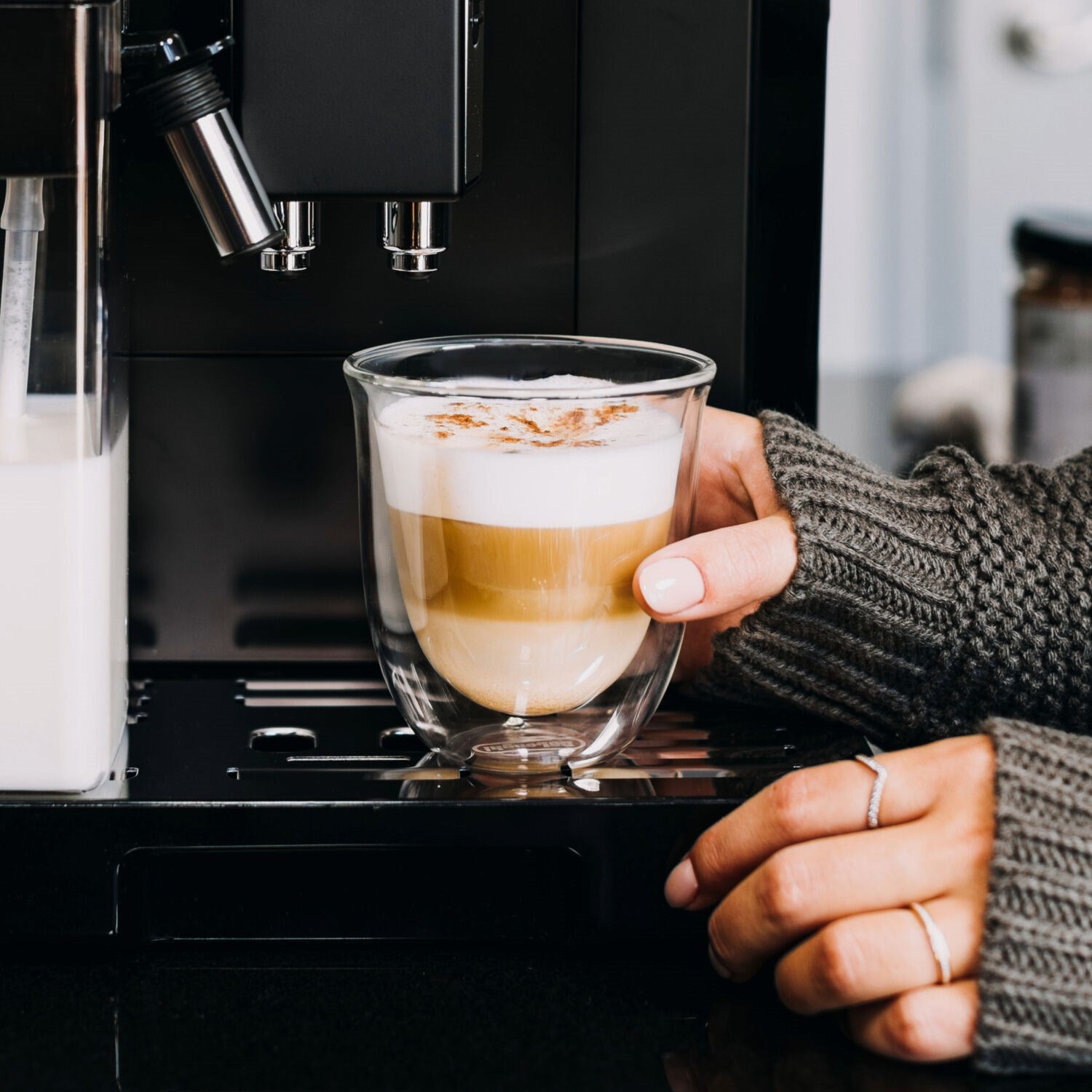 Double-wall clear glass cappuccino cup with layered espresso and foam, held in hand