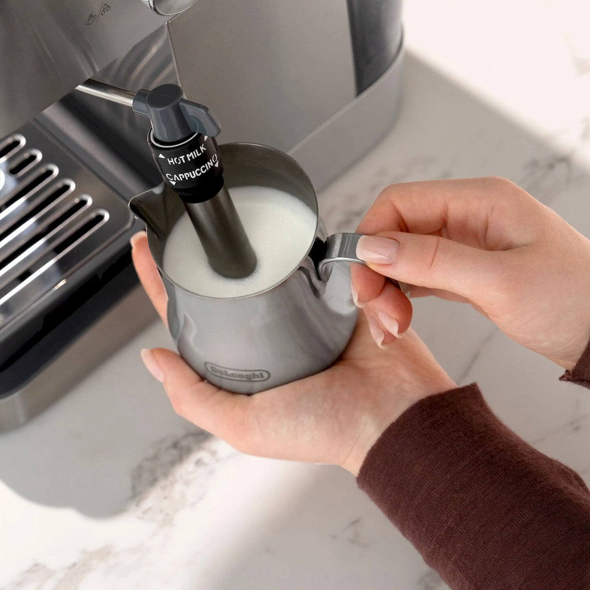 Milk frothing pitcher held under steam wand on stainless espresso machine