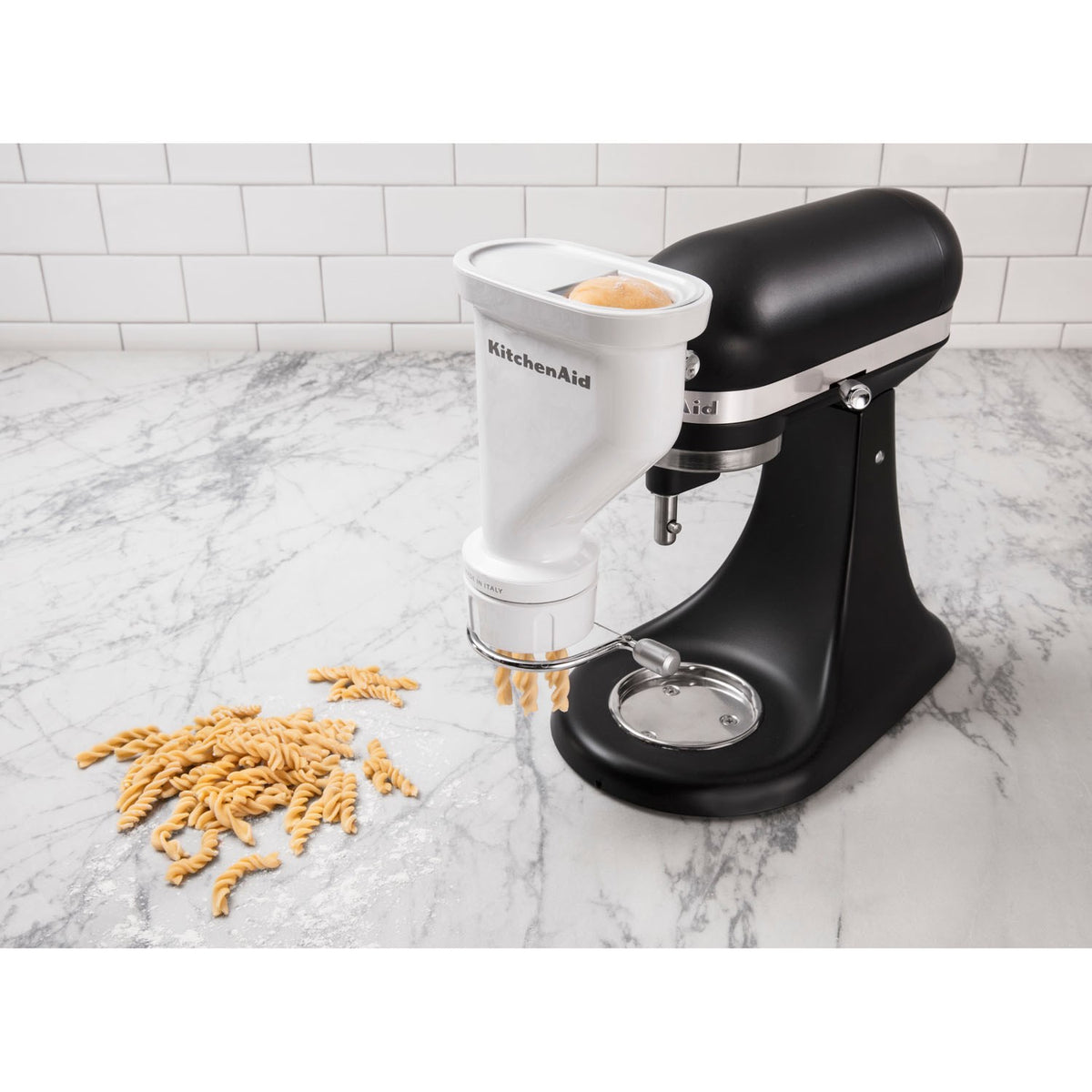 White KitchenAid pasta press attachment mounted on a mixer, with pasta extruding from the die.