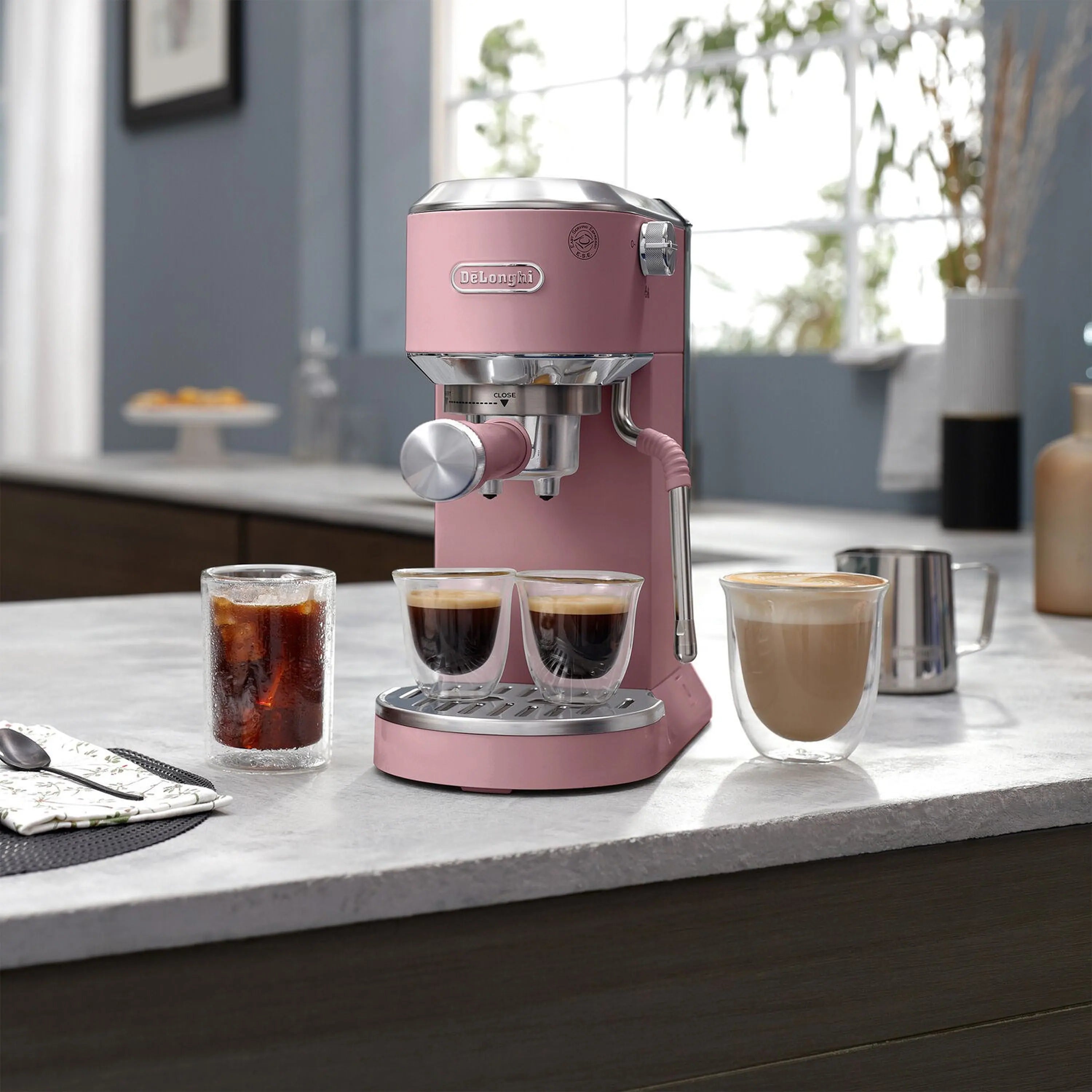 Pink De'Longhi espresso machine with frothing wand, on counter with three coffee cups