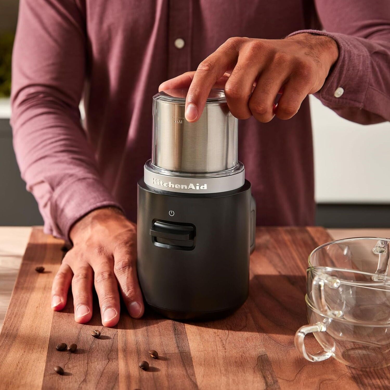 Black coffee grinder on a wooden counter with hand placing whole coffee beans into the lid