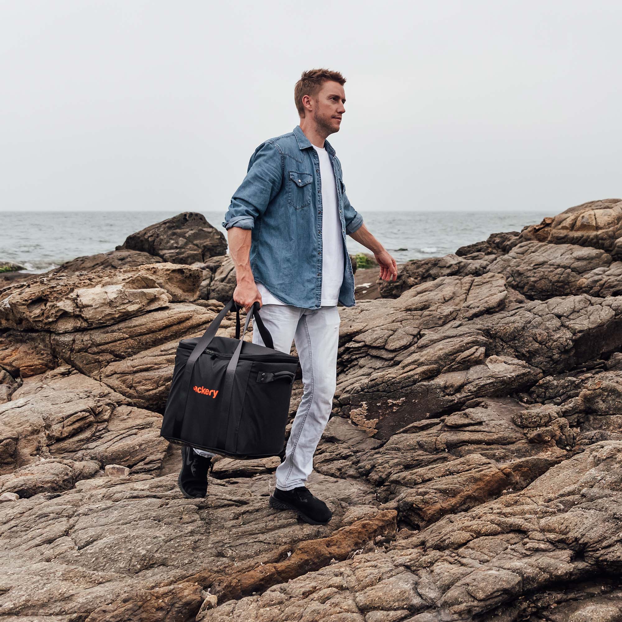 Black Jackery carrying case worn by a person on rocky shoreline, with shoulder strap