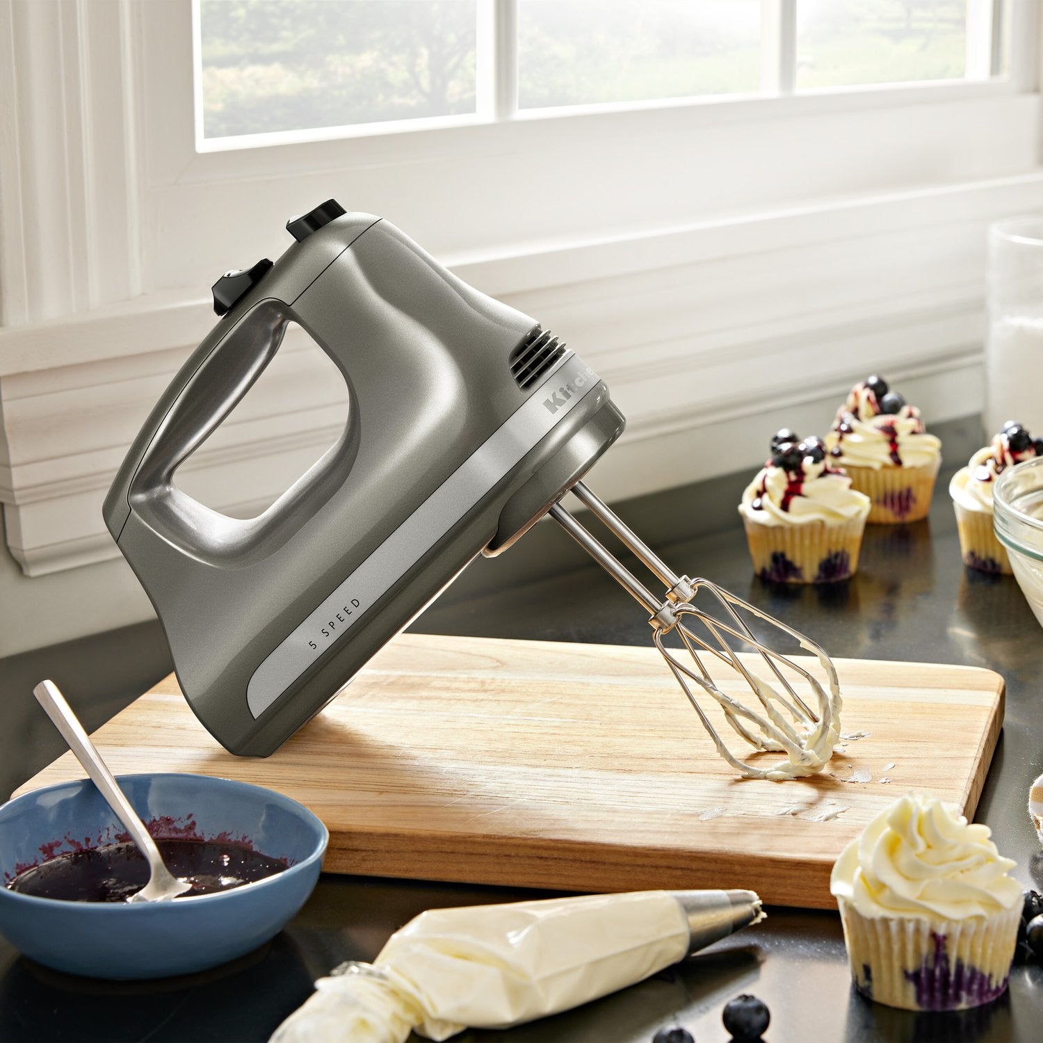 Silver hand mixer with wire whisk beaters in a kitchen counter setting near cupcakes