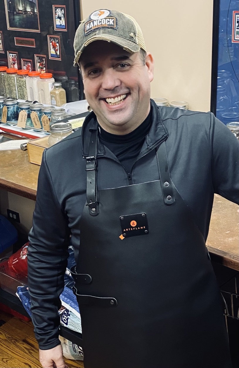 Black leather grill apron with chest logo badge, neck strap, and metal buttons