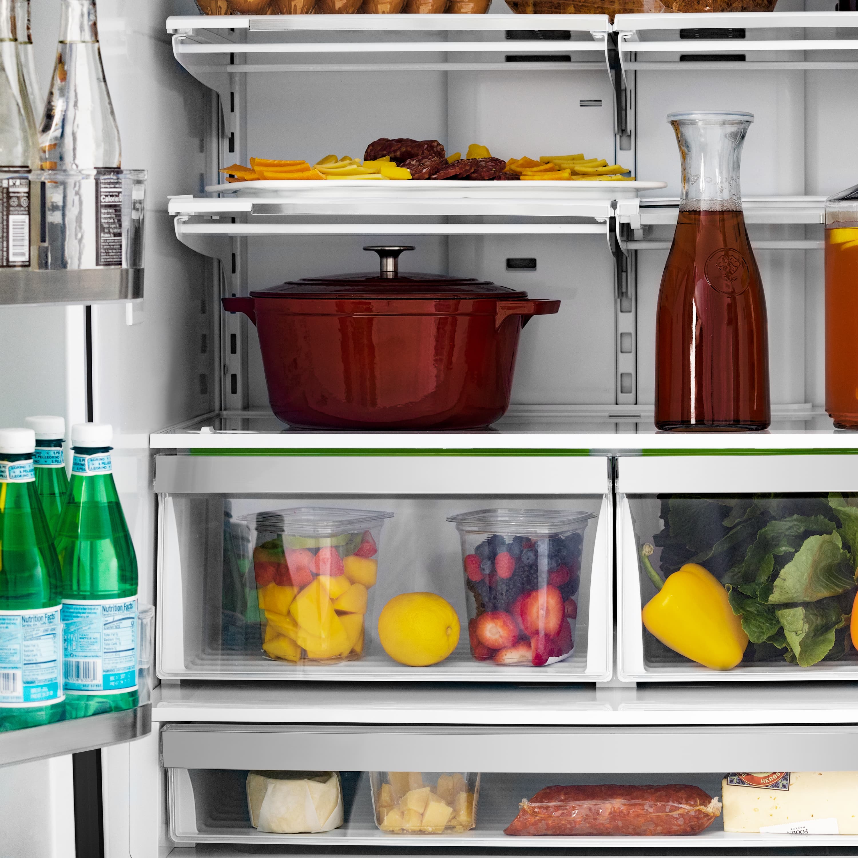 Open refrigerator shelves with a red pot, cheese, vegetables, and drink bottles