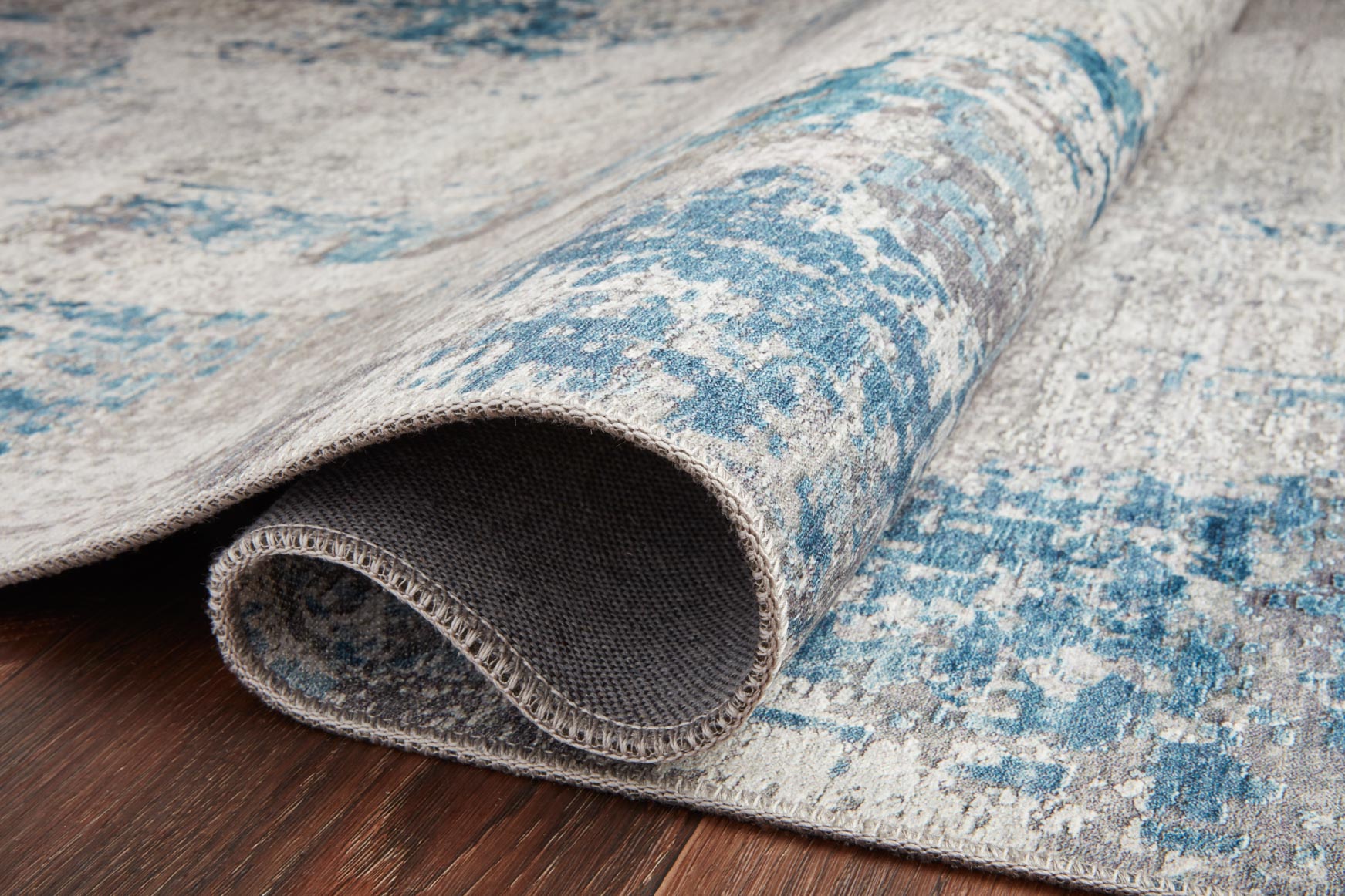 Rolled edge of light gray rug with blue distressed pattern and stitched backing