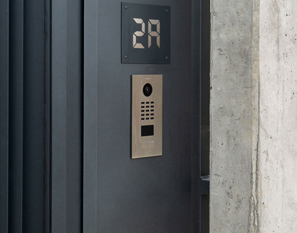 Dark metal video door station panel with camera, keypad, and display.