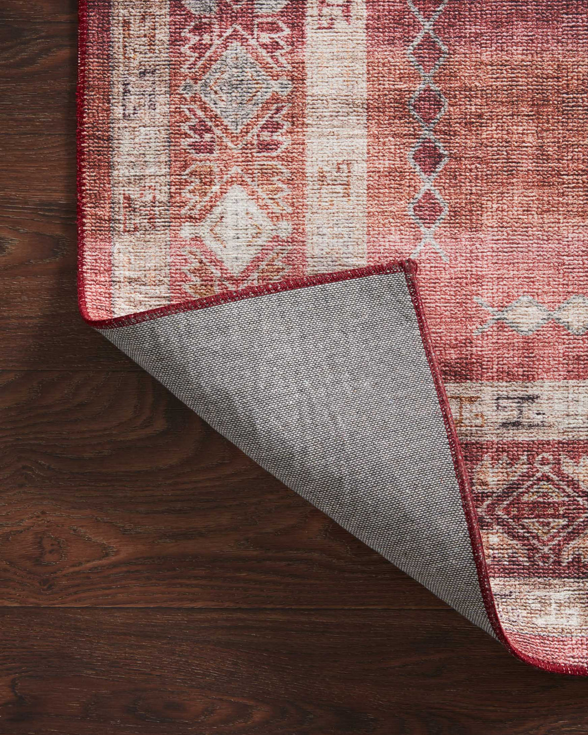 Corner detail of rust red rug with ivory pattern and grey backing visible.