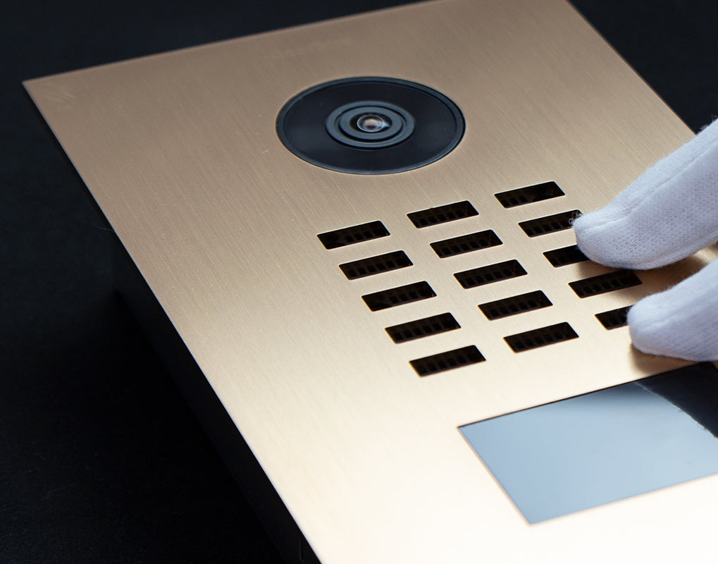 Gold IP video door station with camera lens, keypad grille, and gloved hand