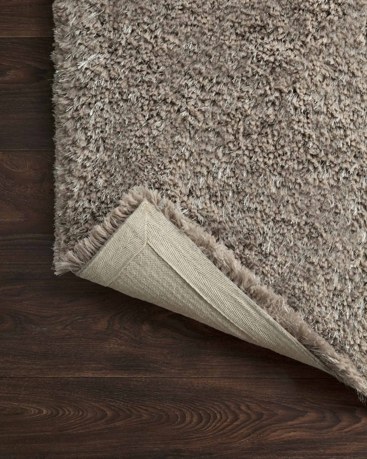 Taupe shag rug corner with visible light backing and stitched seam.