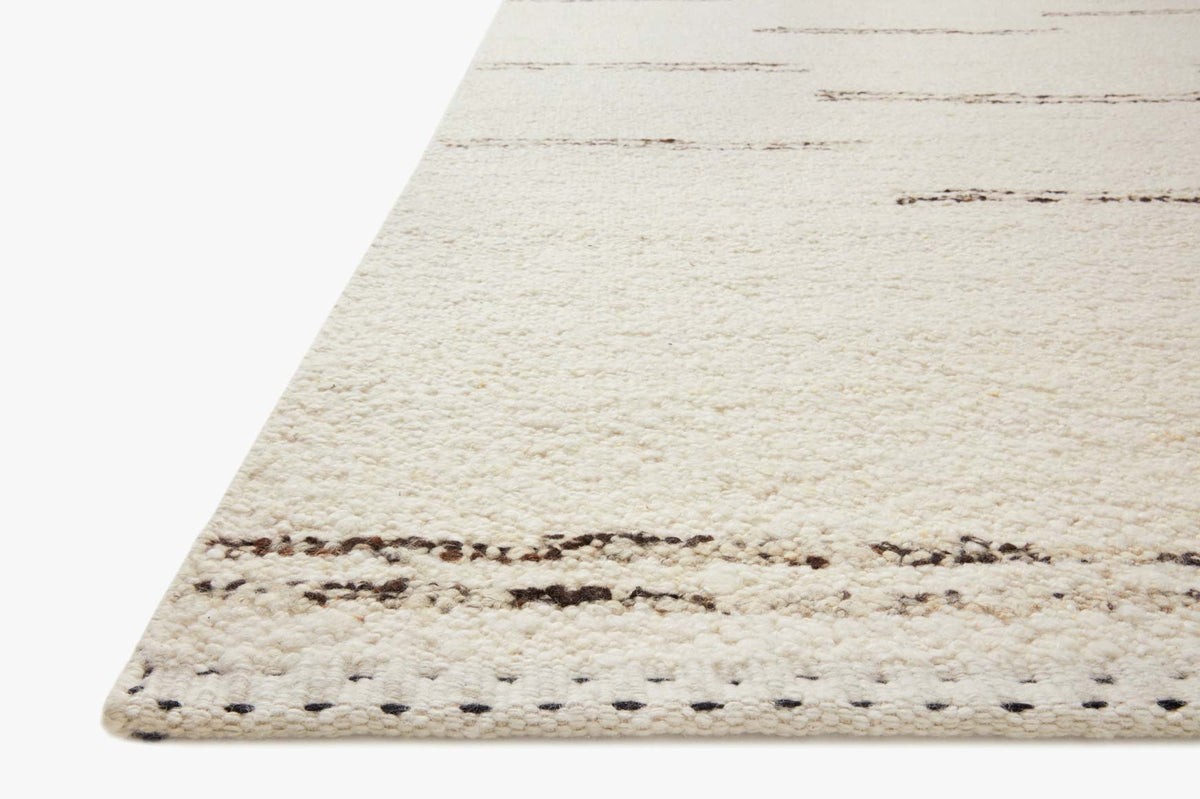 Ivory rug corner with subtle gray-brown edge stitching and fringe detail