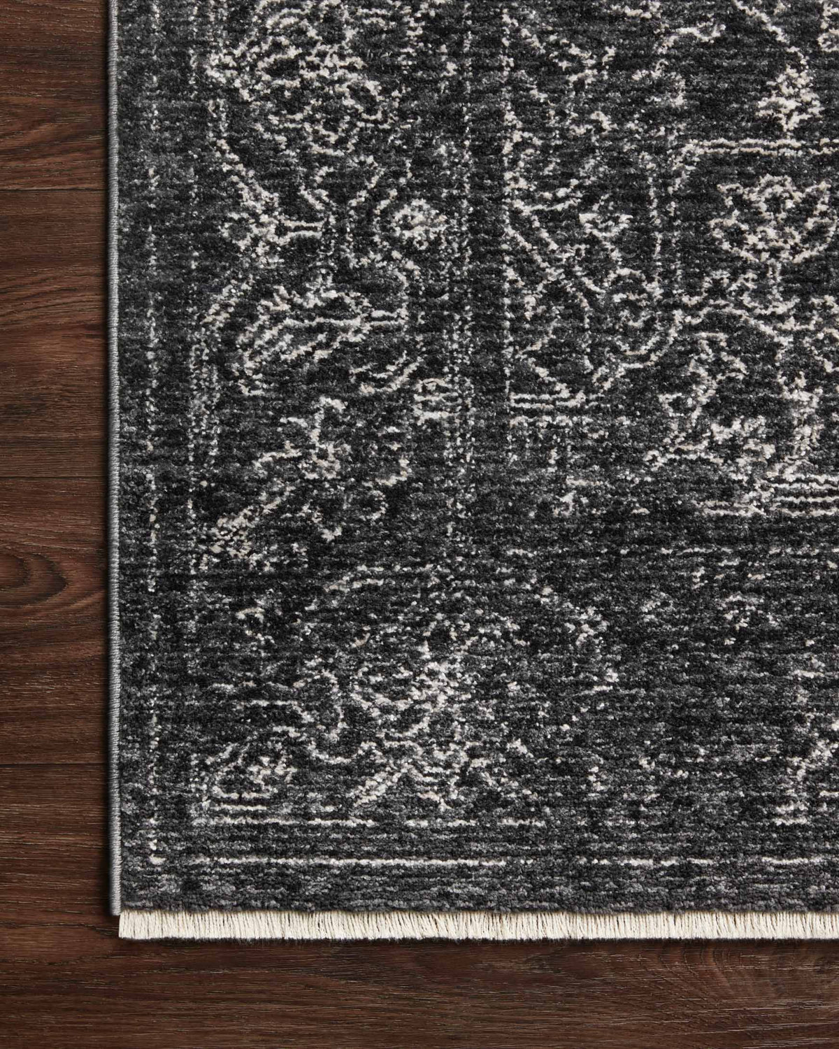 Corner of charcoal and dove rug with white fringe border on dark wood floor.