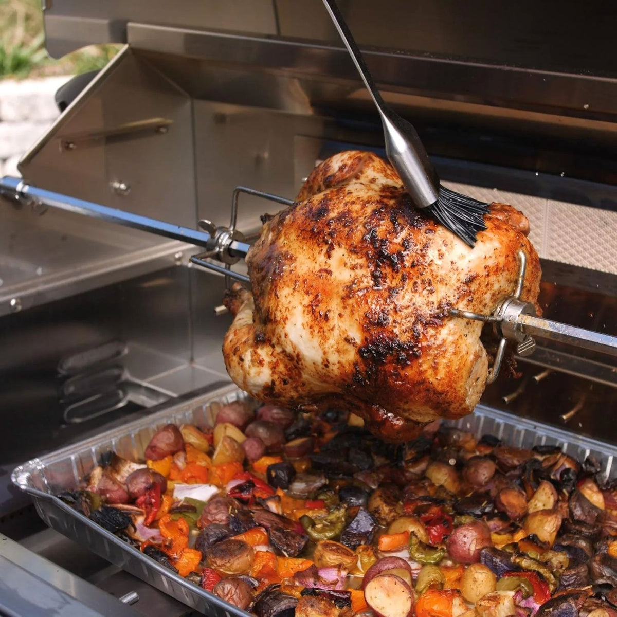 Rotisserie spit with roasted chicken over a drip tray of vegetables
