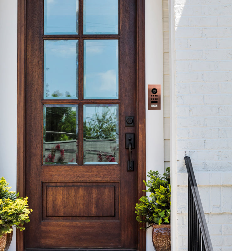 Bronze outdoor video intercom mounted on wall beside door, with call keypad and camera