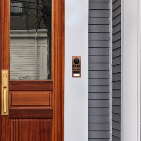 Bronze DoorBird surface-mount video intercom on door frame with camera and call label