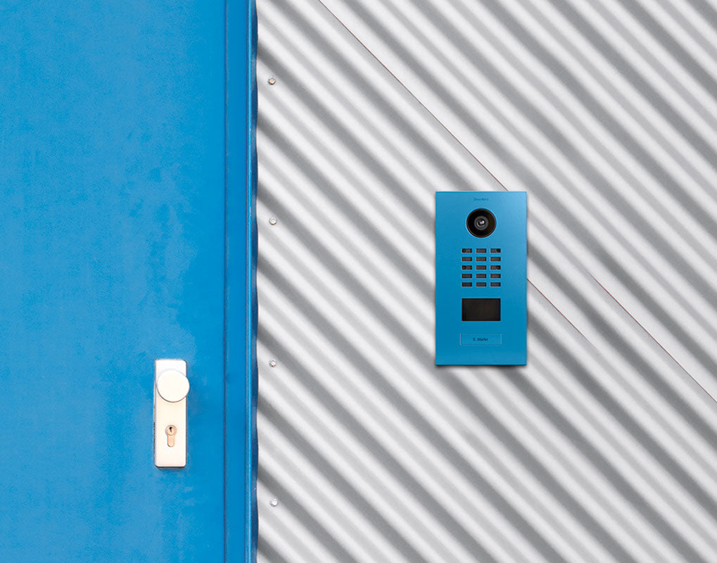 Light blue DoorBird IP video door station on wall with camera and call buttons