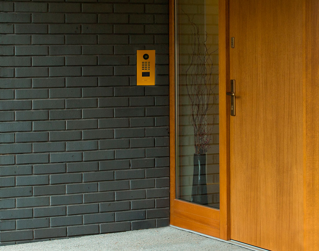 Signal yellow DoorBird IP video door station mounted on a brick wall by a door
