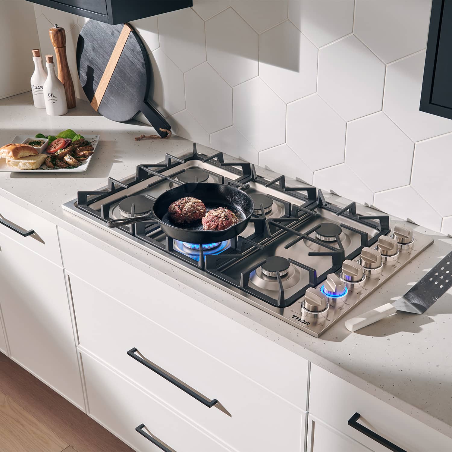 Stainless gas cooktop with black grates; pan cooking on a lit burner with knobs